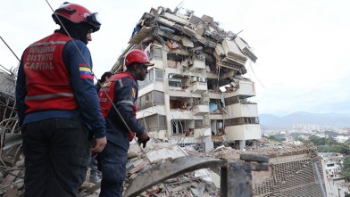 Two firefighters in red helmets and Bomberos Distrito Capital vests look up at a partially collapsed apartment building in Caracas, Venezuela, after the earthquakes.