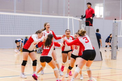 A group of six female volleyball players in red and white uniforms celebrate together on the court, with a volleyball net in the background.