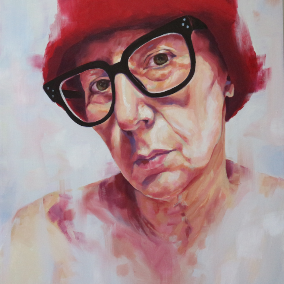 A close-up portrait of a person with short hair wearing a red hat and large black glasses, set against a soft, abstract background.