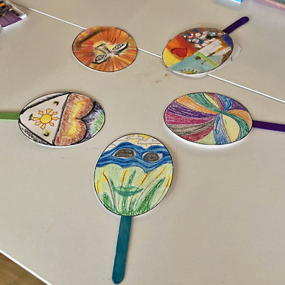 Colorful, hand-drawn circular decorations on sticks arranged on a table, featuring various designs like a sun, hearts, and abstract patterns.