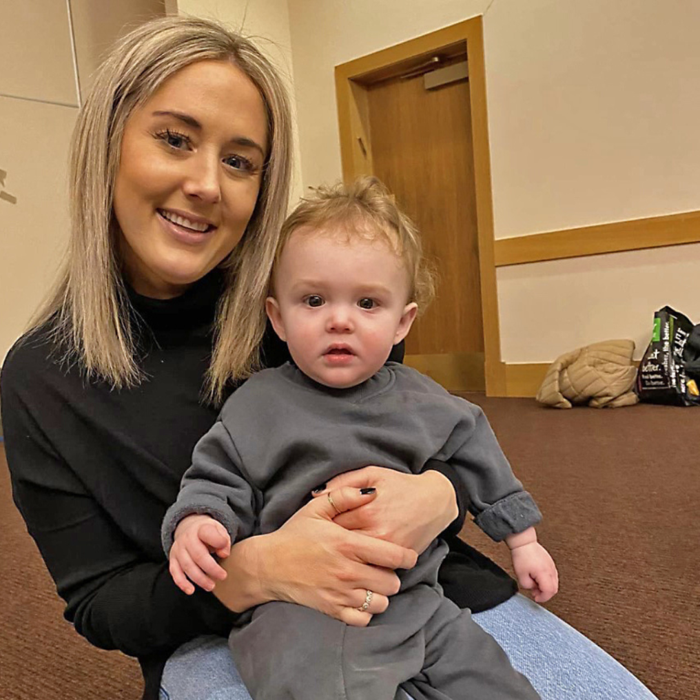 A woman with long blonde hair is sitting on the floor, holding a toddler in gray clothing. They are in a room with a wooden door and a bag in the background.