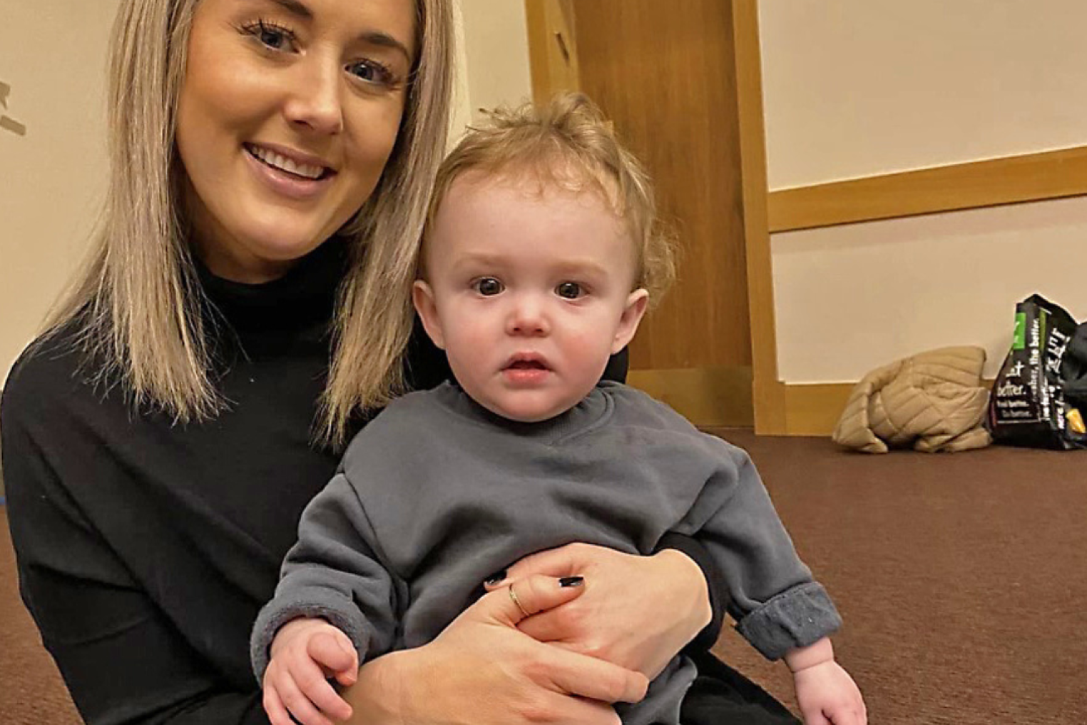 A woman with long blonde hair is sitting on the floor, holding a toddler in gray clothing. They are in a room with a wooden door and a bag in the background.