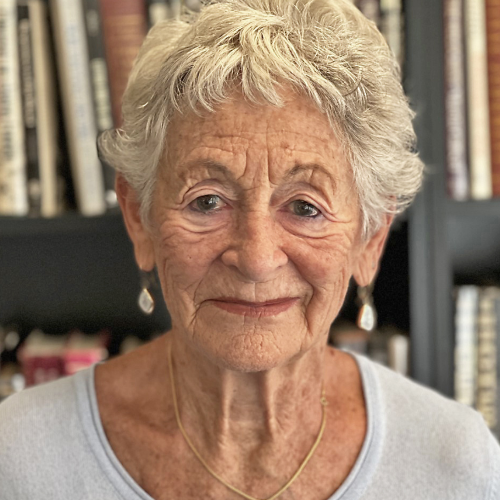 A senior woman with short, wavy gray hair and earrings, smiling softly in front of a bookshelf filled with books.