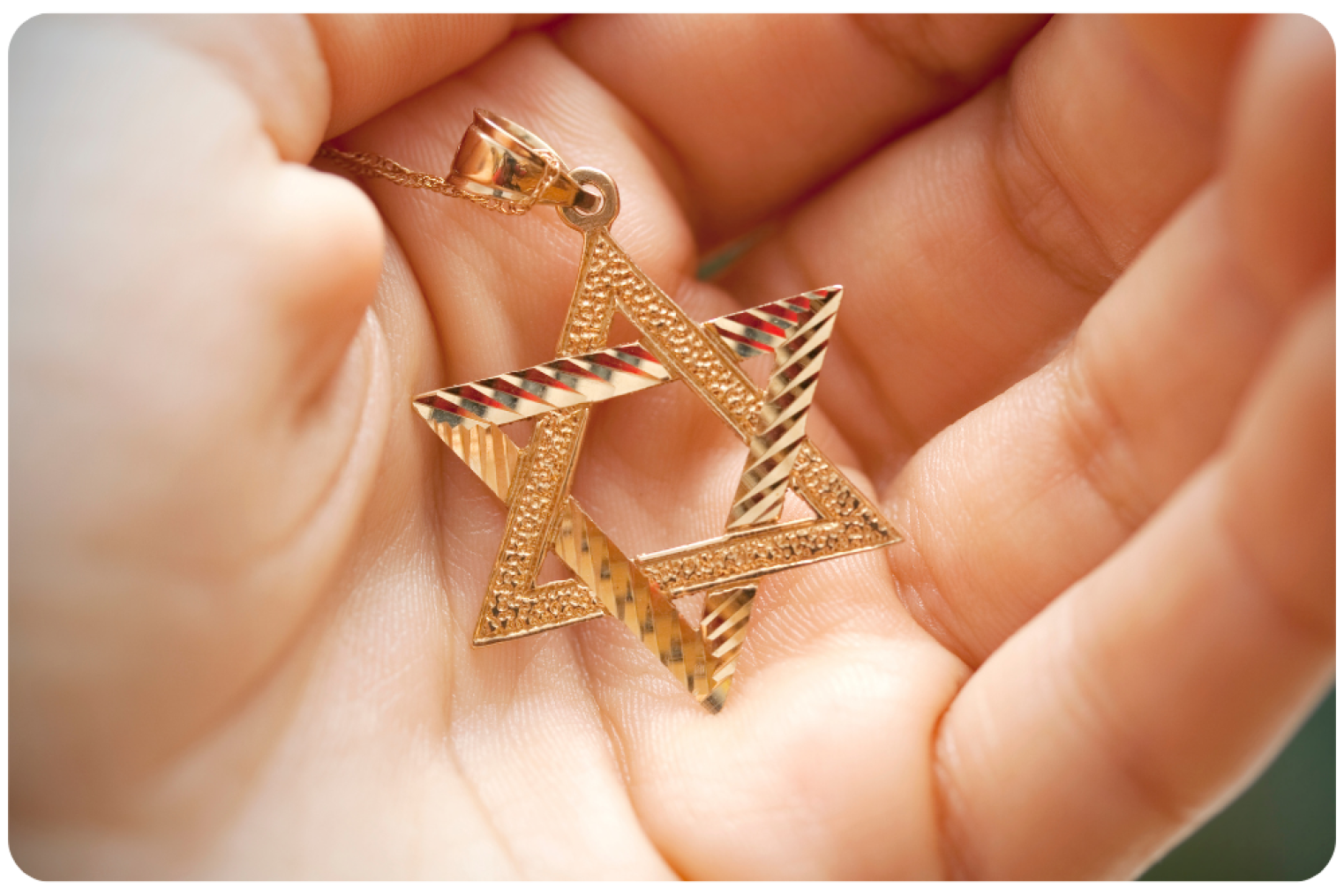 A hand holding a gold Star of David pendant with intricate designs, resting against a soft background.