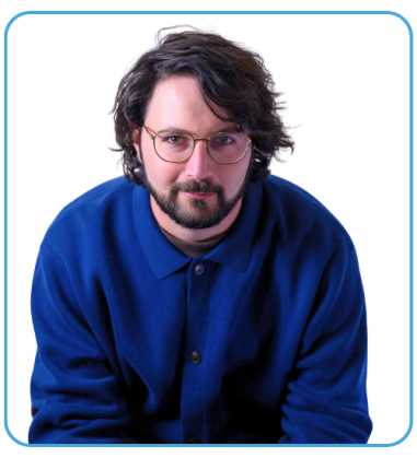 A man with curly hair and glasses wearing a blue button-up shirt is sitting and looking directly at the camera against a white background.