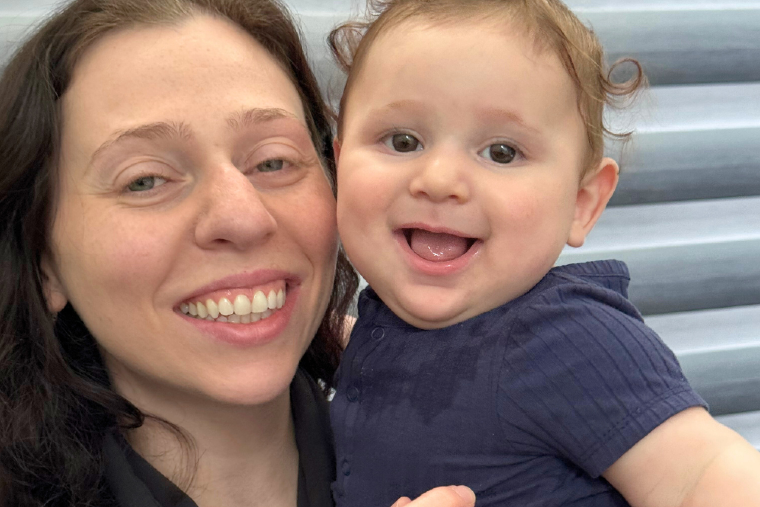 A woman with dark hair smiles while holding a smiling baby with curly hair, both facing the camera against a gray background.