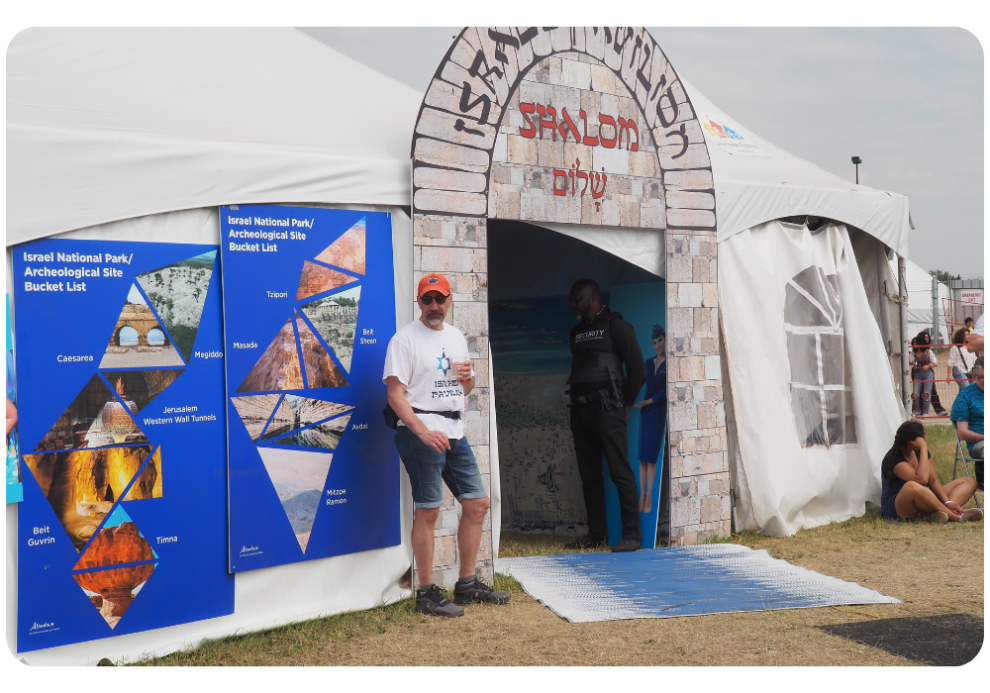 A man in a white shirt stands outside the Israel Pavilion tent beside an archway labeled “Shalom” in Hebrew and English, next to informational panels about Israeli archaeological sites.
