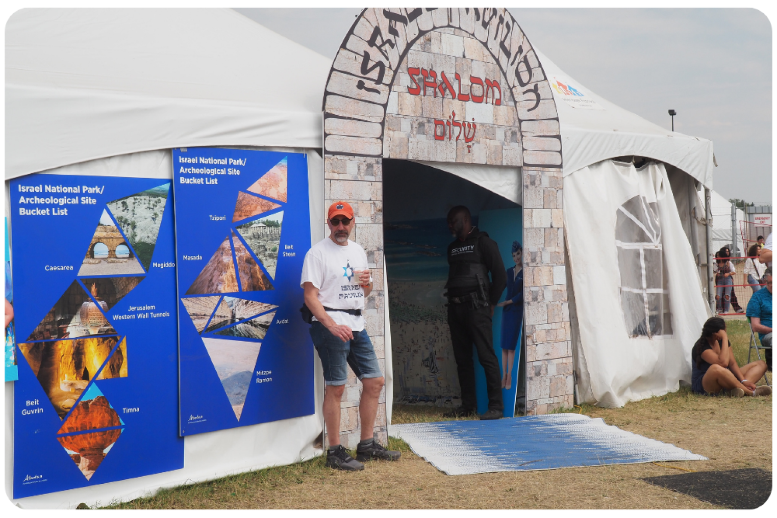 A man in a white shirt stands outside the Israel Pavilion tent beside an archway labeled “Shalom” in Hebrew and English, next to informational panels about Israeli archaeological sites.
