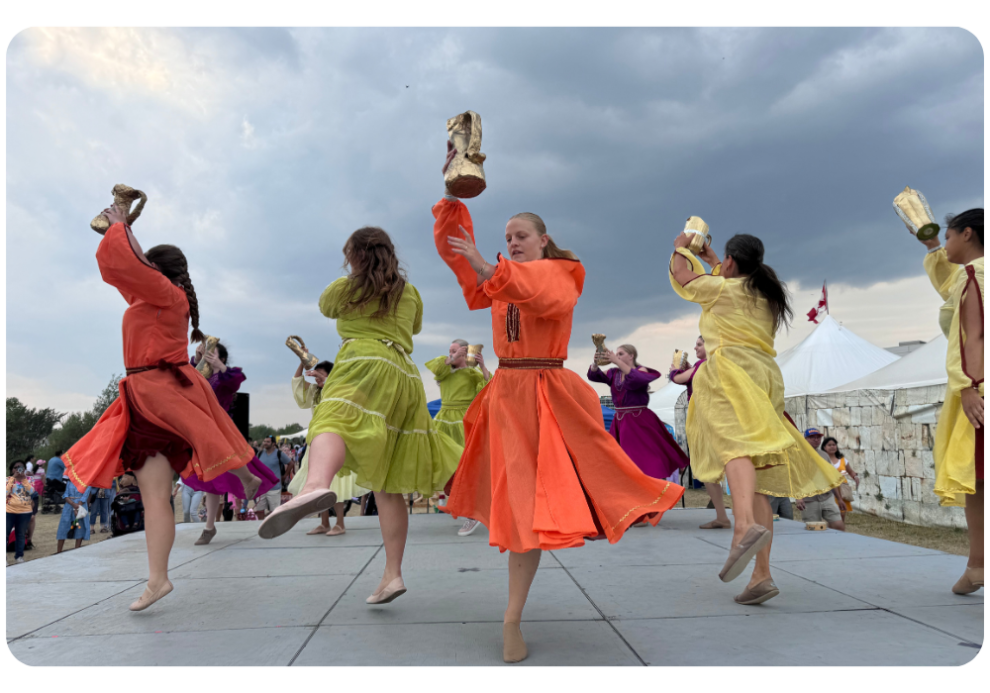 A group of dancers in colorful dresses performing on a stage, holding up objects, with a cloudy sky in the background.