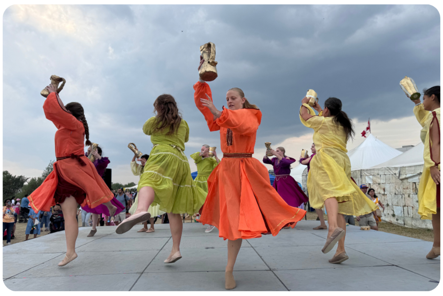 A group of dancers in colorful dresses performing on a stage, holding up objects, with a cloudy sky in the background.