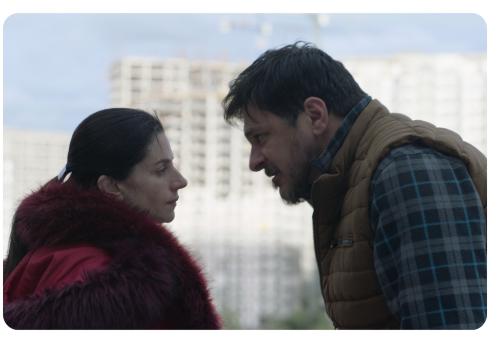 A woman in a red coat and a man in a plaid shirt and vest face each other, appearing to have an intense conversation, with a construction site in the background.