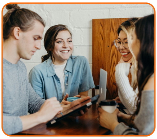Four young adults are gathered around a table, engaging in conversation. One person is using a tablet, while another holds a coffee cup. A laptop is open on the table.