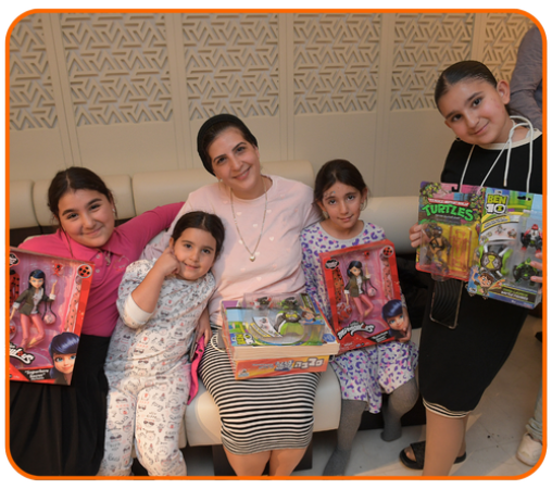 A woman and four girls are seated on a couch, each holding various toys, including dolls and action figures. The background features a decorative wall.