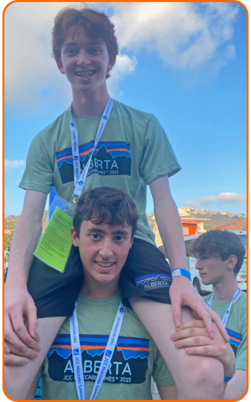 Two boys wearing matching green shirts with "ALBERTA" and "JCC Maccabi Games 2023" are posing outdoors. One boy is sitting on the other's shoulders, both smiling. A third boy is partially visible in t
