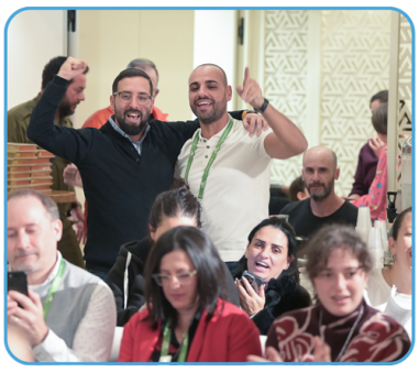 Two men stand with raised arms, celebrating, in a crowded room filled with seated attendees. Several people in the audience are smiling and looking towards the front.