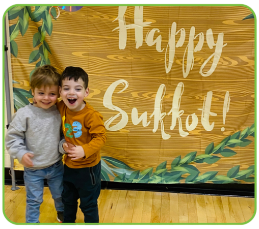 Two young boys stand together, smiling in front of a wooden backdrop that says "Happy Sukkot!" with green leaves.