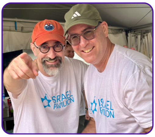 Two men smiling and posing for the camera, wearing white shirts with "ISRAEL PAVILION" printed on them. One man has a red baseball cap and glasses, while the other wears a green cap and glasses.