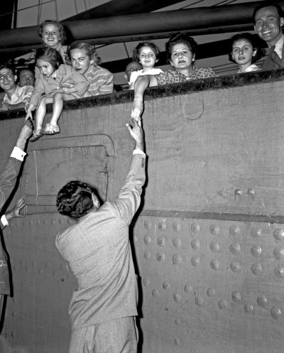 A group of people, including children, are gathered on a ship's deck, reaching out to a man in a suit below who is extending his hand towards them.