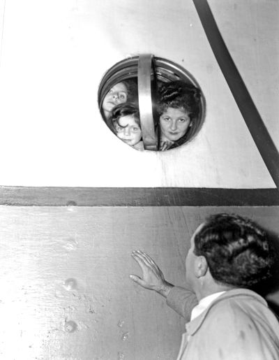 Three children peering through a round porthole on a ship, while a man reaches out towards them from outside.