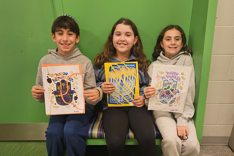 Three children sitting on a bench against a green wall, each holding colorful artwork featuring Jewish symbols