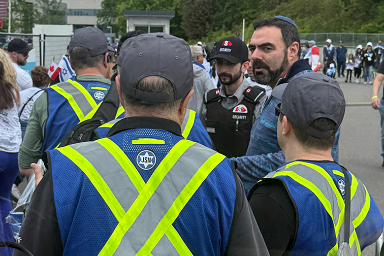 A group of people wearing blue vests with reflective yellow stripes, some with caps, are gathered near a security personnel in a black uniform, outdoors at an event