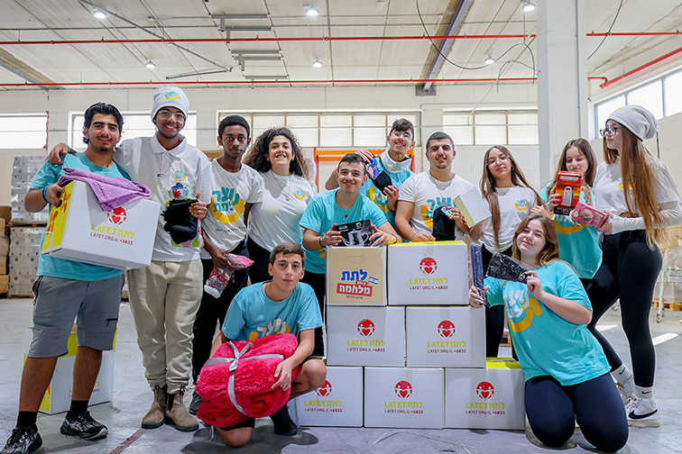 A group of ten young people in matching t-shirts poses in a warehouse, holding various items and standing in front of boxes labeled