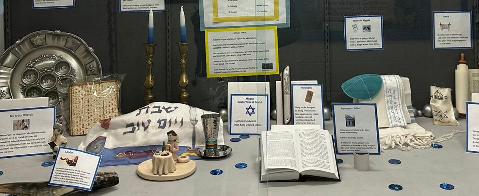 A display case featuring various Jewish ritual items, including a Seder plate, menorah, Torah, and Shofar, with informational labels.
