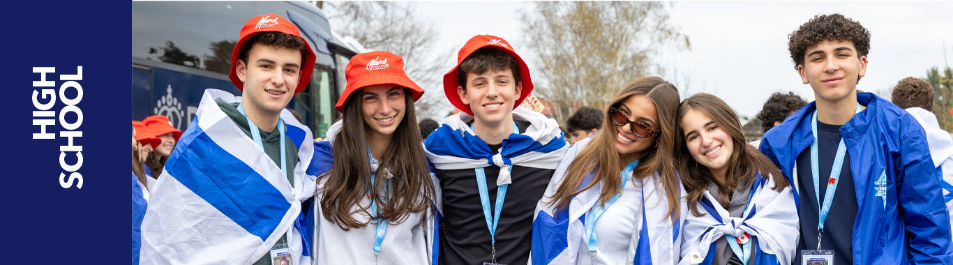 A group of high school students, wearing blue and white clothing and holding Israeli flags, pose together outdoors, smiling. 