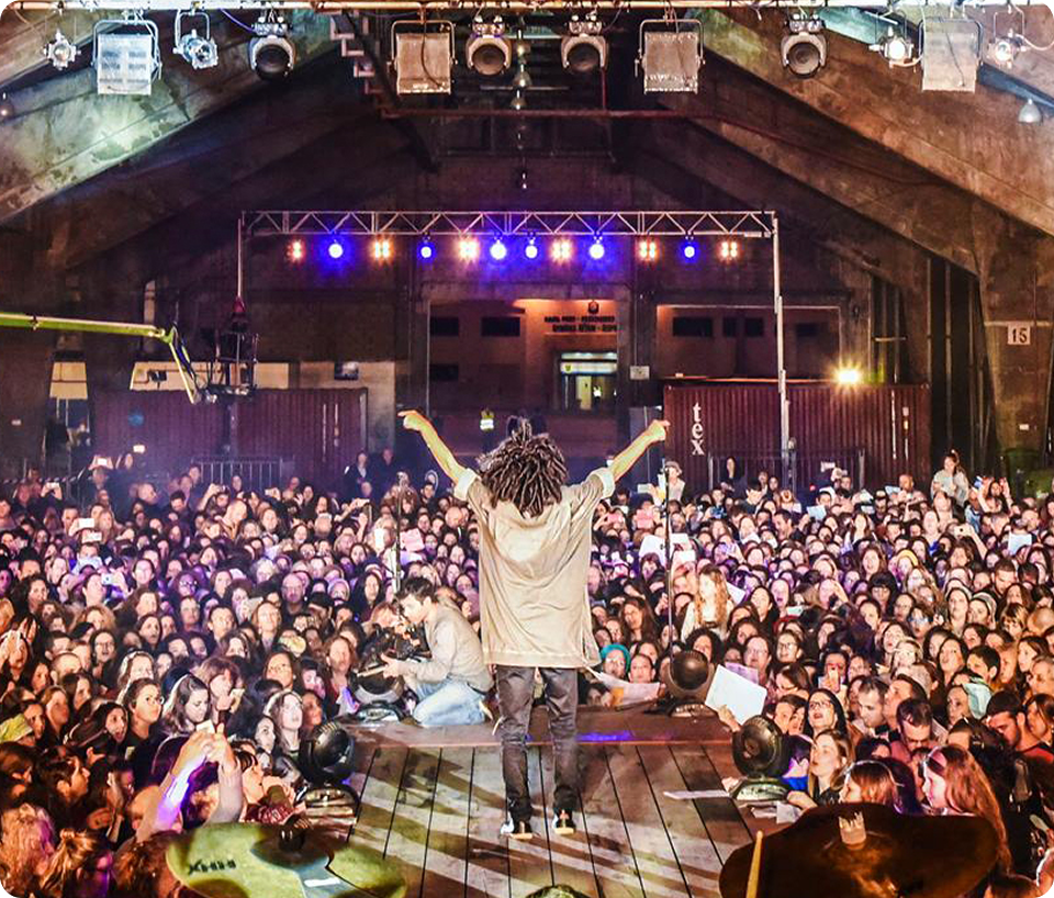 A performer with curly hair stands on stage, facing a large crowd of fans in a dimly lit venue with colorful lights.