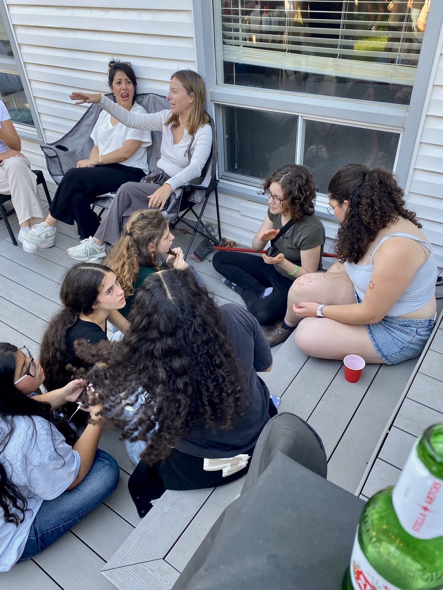 A group of young women sitting on a wooden deck, engaged in conversation; two women are seated in chairs, while others sit on the ground. A red cup and a beer bottle are visible in the foreground.