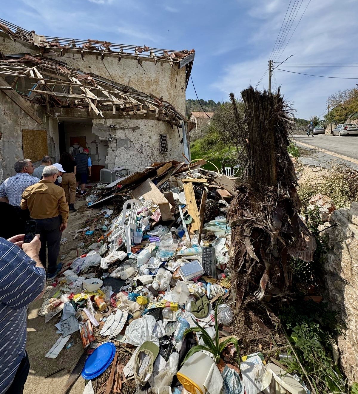 A group of people standing near a damaged building with a partially collapsed roof, surrounded by debris and trash on the ground.